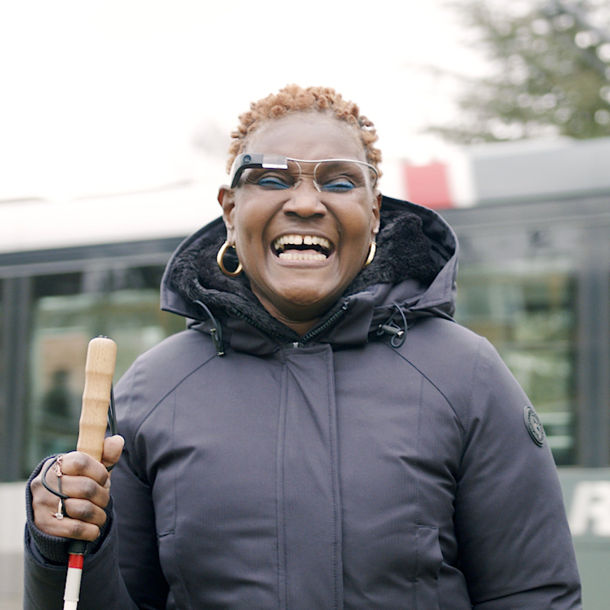 A photo of Joy who's wearing the Envision Glasses with the titanium frames and holding a white cane on her right hand while facing towards the camera.